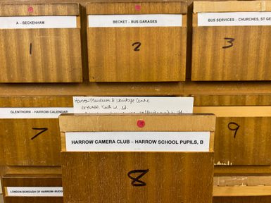 A set of wooden archive card catalogue drawers arranged in rows. One drawer is pulled open and labelled “Harrow Camera Club – Harrow School Pupils, B,” with a handwritten index card partially visible inside. The surrounding drawers are numbered and labelled with other Harrow history subjects.