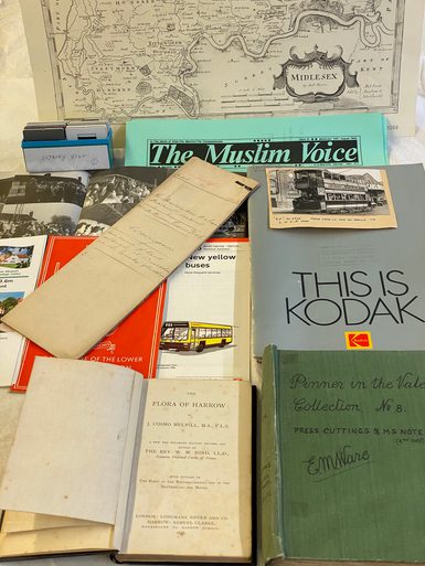 A display of books, photographs, and printed materials from the Harrow archives arranged on a table. Items include The Muslim Voice newspaper, a booklet titled This is Kodak, a bus leaflet about new yellow buses on route 258, and an open copy of The Flora of Harrow (1876). A scrapbook titled Pinner in the Vale Collection and a historic map of Middlesex are visible in the background.