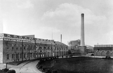 Black-and-white historic photograph of the Kodak factory complex in Harrow. A large brick industrial building in the foreground displays prominent “KODAK” signage along the roofline and across the façade, with additional wording referencing Eastman and photo materials. A tall chimney rises from the site, with light smoke visible. The buildings surround a curved driveway and landscaped lawn, with a few small figures walking near the entrance.