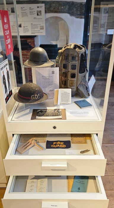 A museum display case featuring Second World War home front objects, including helmets, a gas mask, documents and photographs. Open drawers beneath the case contain additional items such as a “Fire Guard” armband, air raid information leaflets and small tools used for wartime civil defence.