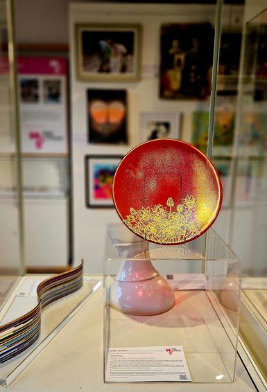 A red glass decorative plate with gold floral detailing displayed on a stand inside a clear museum case. The plate forms part of an exhibition display, with colourful glass samples and framed artworks visible in the softly lit gallery space behind it.