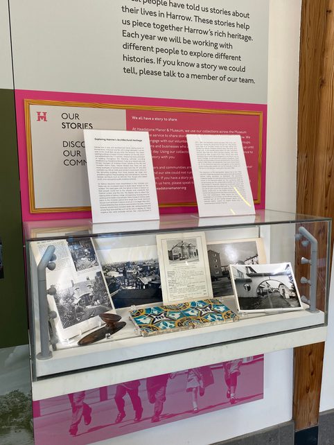 A museum display case featuring photographs and objects relating to Harrow’s architectural heritage. Black-and-white photographs of buildings are arranged alongside a decorative ceramic tile and a small wooden tool, with interpretation panels and a sign titled “Our Stories: Discover Our Community” behind the case.