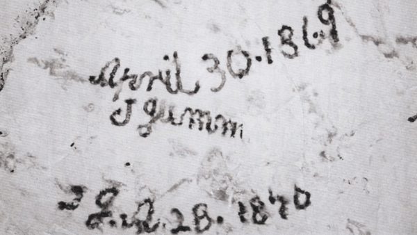 Close-up photograph of handwritten chalk markings on a pale stone wall inside a chalk mine, showing historical signatures left by workers or visitors.