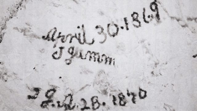 Close-up photograph of handwritten chalk markings on a pale stone wall inside a chalk mine, showing historical signatures left by workers or visitors.