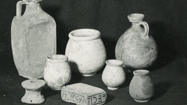 Black-and-white photograph of several weathered ceramic vessels and a carved rectangular artifact displayed against a dark background, likely part of an archaeological collection.