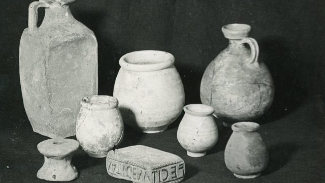 Black-and-white photograph of several weathered ceramic vessels and a carved rectangular artifact displayed against a dark background, likely part of an archaeological collection.