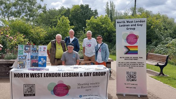 A photograph of 5 members of the North West London Lesbian and Gay Group sat at a stall on Headstone Manor and Museum's grounds. The stall is filled with leaflets. Next to it sits a pop up banner with a LGBTQIA+ flag printed on the front.