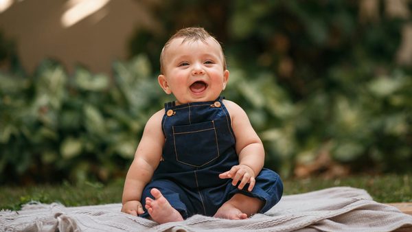 A baby wearing denim dungarees sat on a blanket outdoors
