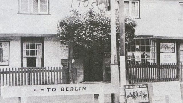 Black and white photograph of large house facade. 1705 on its face, with flowers above the doorway and a sign pointing left 'To Berlin.'