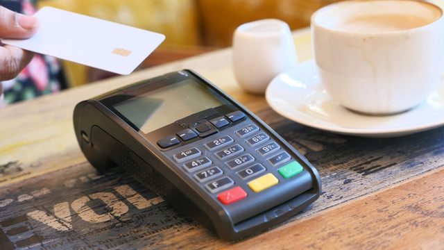 Hand placing bank card on card reader laid on a counter, paying for a coffee which sits beside the machine on the counter