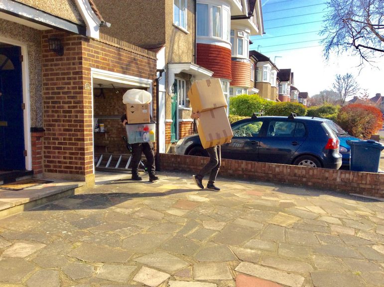 Two people carrying moving boxes from a garage of a house