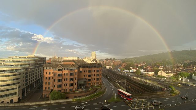 Rainbow arching over road, buildings on one side, houses and green hill on the other side