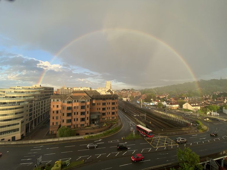 Rainbow arching over road, buildings on one side, houses and green hill on the other side