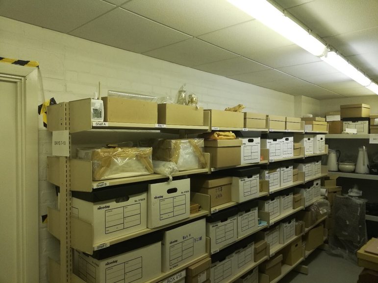 Storage facility for the collection at Headstone Manor and Museum. Cardboard archival boxes stacked on shelves, low lighting.