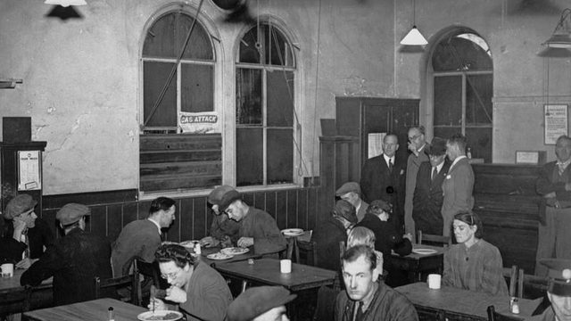 Black and white image of a small dining hall filled with men and women eating
