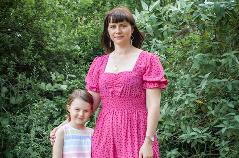 A mother with a pink dress and brown hair and daughter with a stripey pastel shirt stood together in greenery