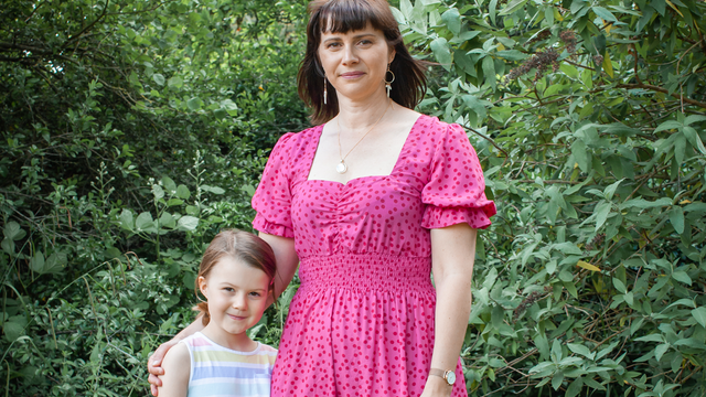 A mother with a pink dress and brown hair and daughter with a stripey pastel shirt stood together in greenery