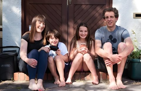 A family of four sat on the doorstep of their house; a mother, son, daughter and father, brown door behind them.