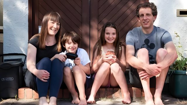 A family of four sat on the doorstep of their house; a mother, son, daughter and father, brown door behind them.