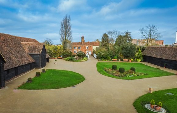 Aerial shot showing the great barn to the left, the manor house in the back and the entrance to the right. Patches of green grass while beige pavement weaves through the ground as a clear and identifiable walking path.
