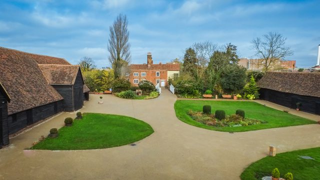 Aerial shot showing the great barn to the left, the manor house in the back and the entrance to the right. Patches of green grass while beige pavement weaves through the ground as a clear and identifiable walking path.