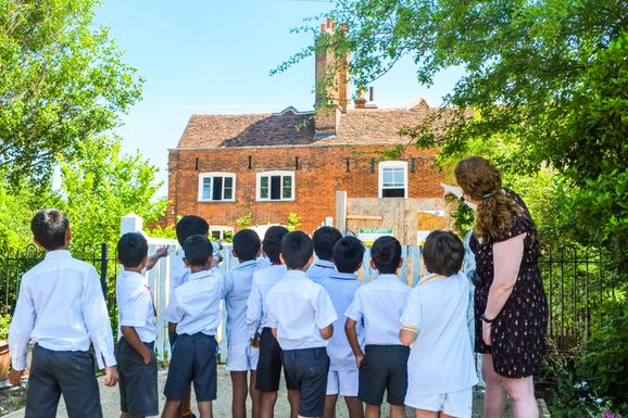 A teacher is pointing towards an old manor house as her students look up. You can only see the back of the heads.