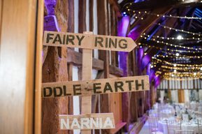 A wooden signpost with "Fairy Ring", "Middle Earth" and "Narnia" is leant against the wooden beams of the barn. Round dining tables, purple uplighters and fairy lights are visible in the background