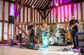 A band performs under purple uplighters with the wooden beams of the Great Barn visible behind them
