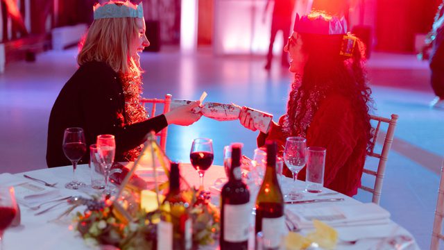 Two women in Christmas hats sit at a dinner table, the lights are dimmed and there is blue and red lighting