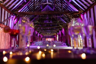 A long shot of the Great Barn with the lights fully dimmed, purple and red uplighters and fairy lights hung from the ceiling beams. In the foreground, champagne and cocktail glasses sit on the bar decorated with fairy lights in soft focus