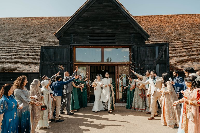 Bride and groom walking through the archway of the great barn beneath confetti thrown by happy wedding guests